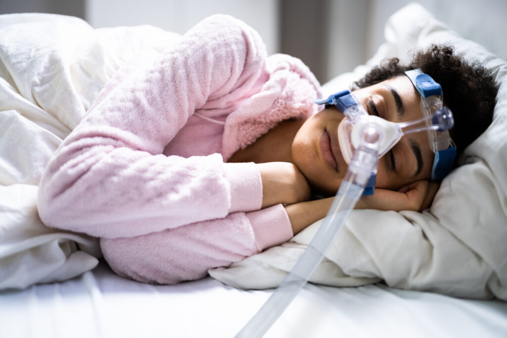 Can You Get a CPAP Machine Without Insurance? Woman in pink sweater with curly hair sleeping with a cpap mask on.